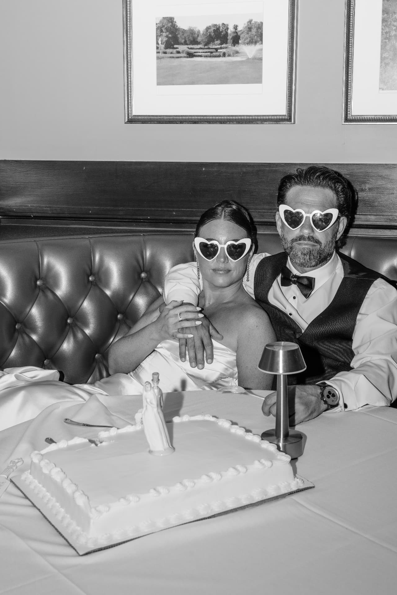 Black and white photo of a bride and groom in heart sunglasses beside their cake