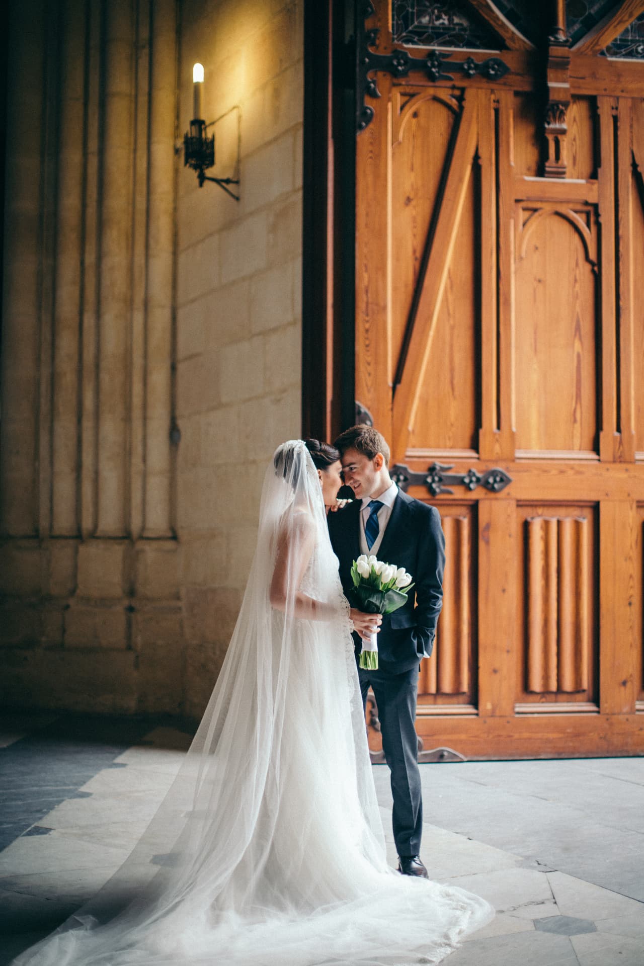 Bride with a long veil and groom touching foreheads beside carved cathedral doors