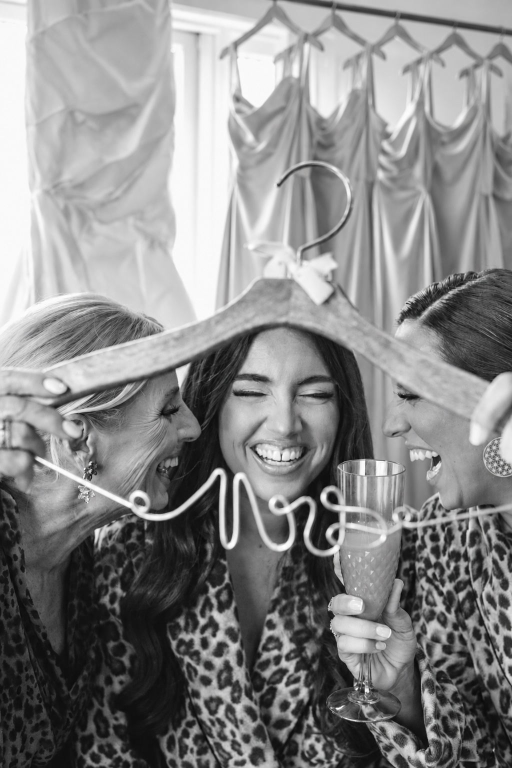 Black and white photo of the bride laughing with her mom and maid of honor behind a Mrs. hanger