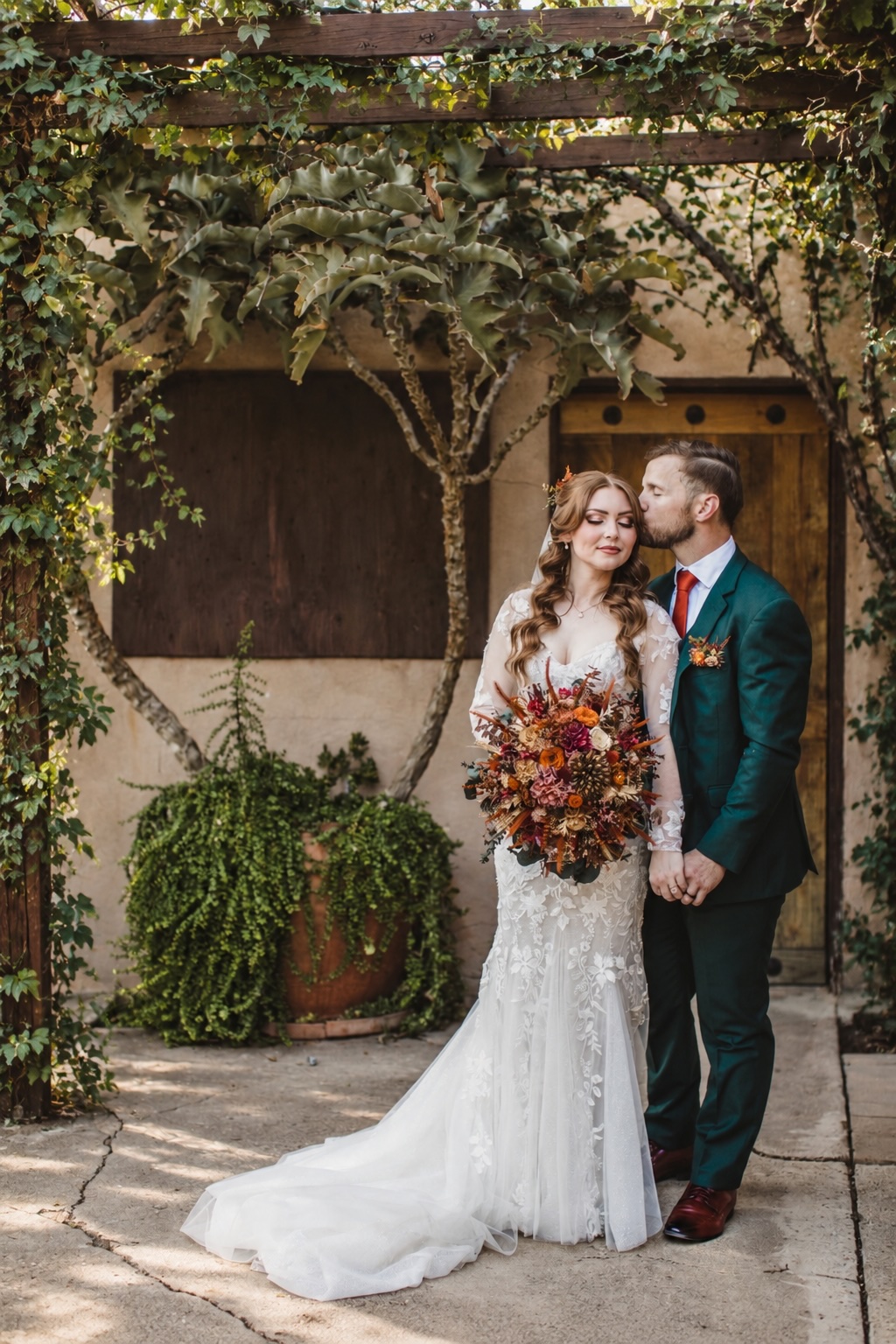 Groom kissing his bride's forehead in an ivy-covered courtyard with a dried floral bouquet