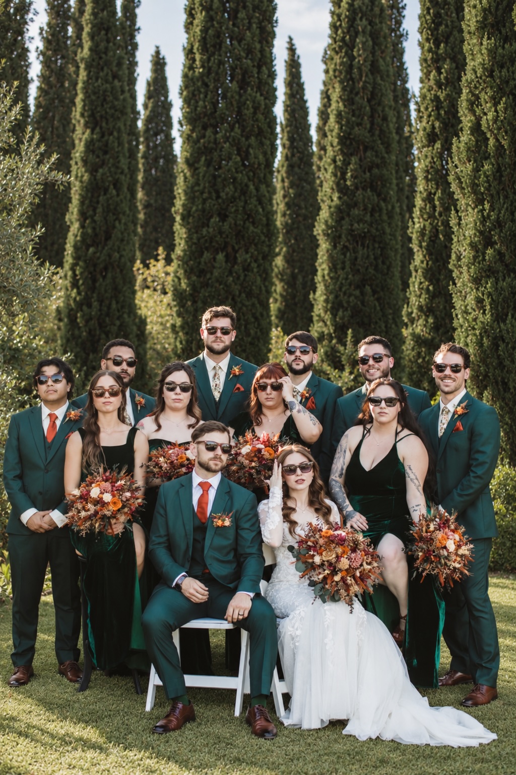 Wedding party in green suits and velvet dresses wearing sunglasses beneath cypress trees