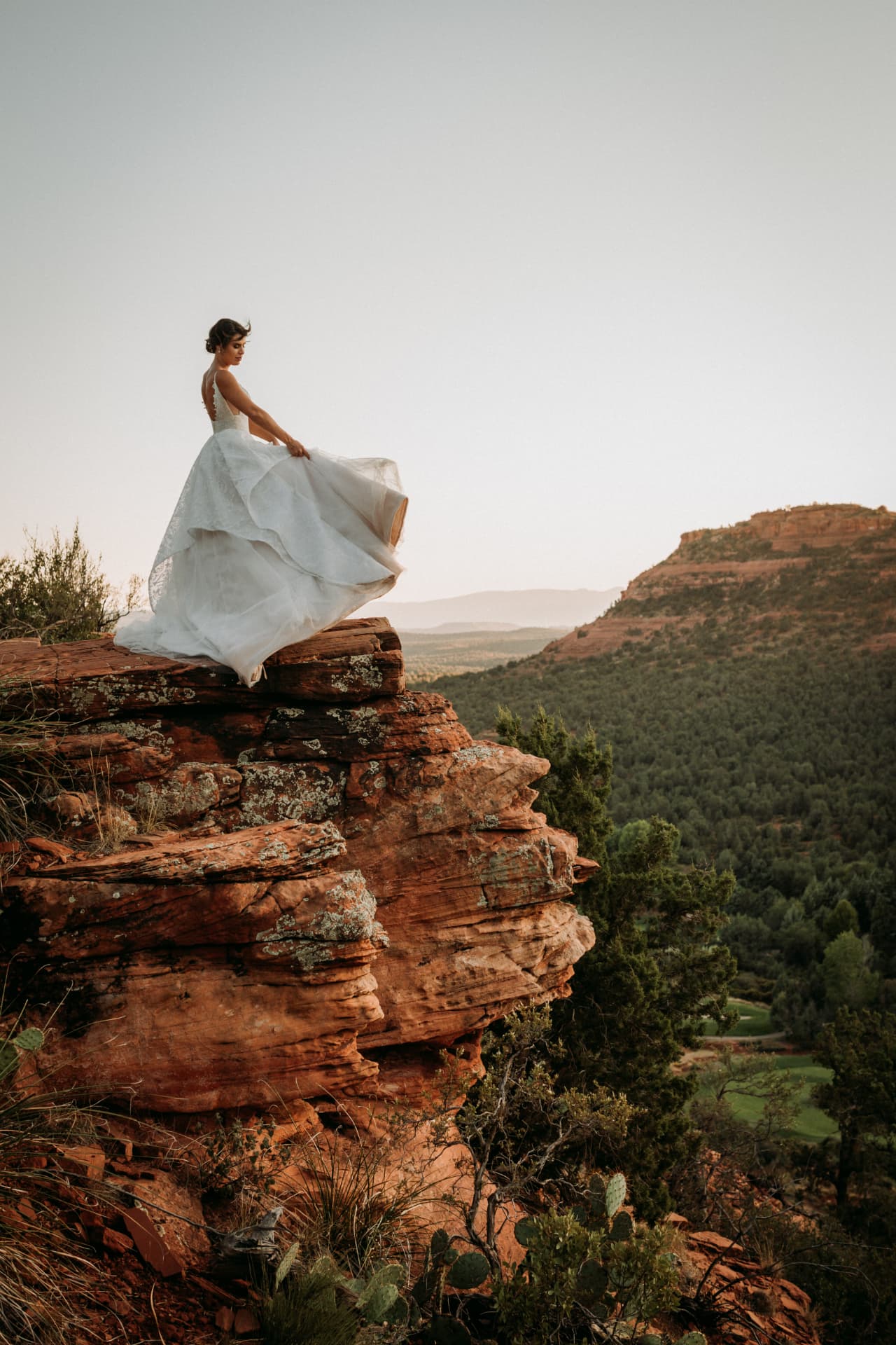 Bride in a flowing white gown standing on red Sedona rocks at sunset